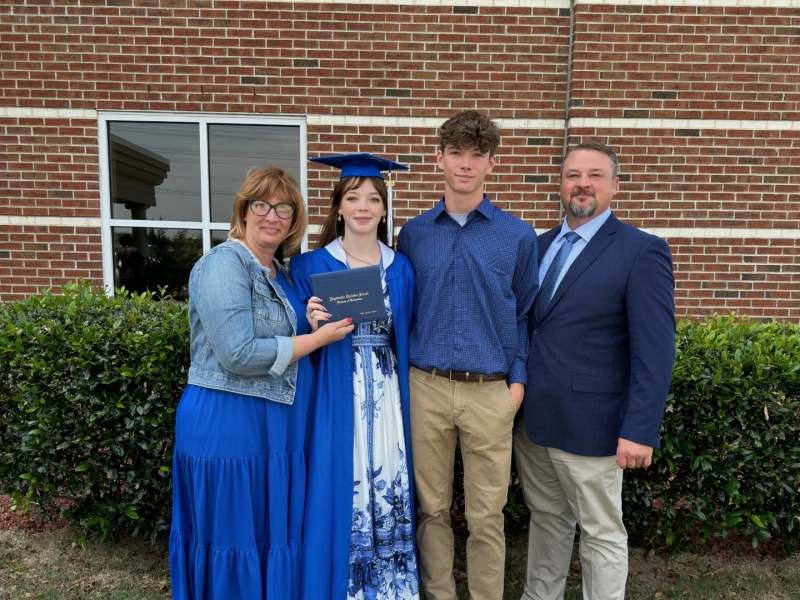 Lilly Guinn stands in blue cap and graduation gown, holding her diploma smiling alongside her mother, father, and brother.